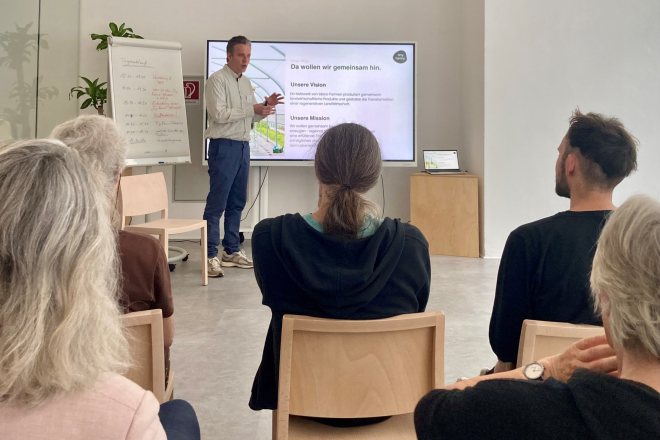 Ein Mann hält eine Präsentation vor einem Bildschirm. Auf der Folie sind die Texte "Unsere Vision, Unsere Mission" zu sehen und daneben steht ein Flipchart mit Notizen. Das Publikum sitzt und hört aufmerksam zu.