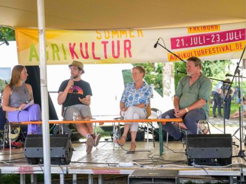 Auf einer Bühne im Freien vor einem Banner des Agrikulturfestivals sitzen von links nach rechts: Moderatorin Sophia McRae (Sustainable Food Economy Lab, Universität Freiburg und Organisationsteam Agrikulturfestival), Sebastian Rogga (ZALF), Sabine Fey (Geschäftsführerin Großmarkt), Wolfgang Hees (Bio-Bauer, Teil EZG Biogemüse Südwest, Projektleiter "Wir bündeln Bio")