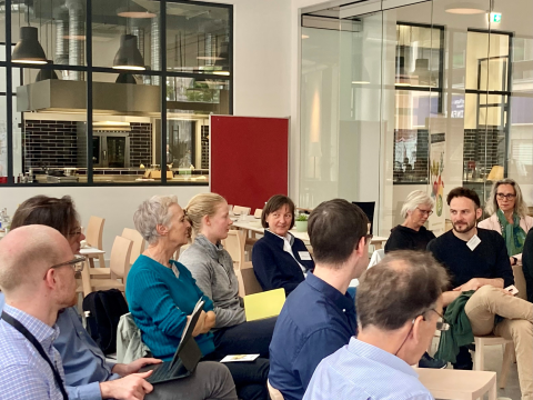 Eine Gruppe von Personen sitzt in einem modernen Konferenzraum mit Glaswänden und Blick in eine Küche. Sie sind in eine Diskussion vertieft, einige halten Notizblöcke und Laptops.